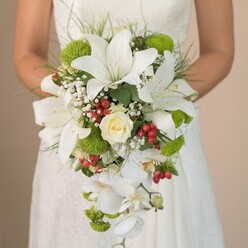 Cradled in the bride's hands against the delicate lace of her wedding gown, this extraordinary cascading bouquet seems to glow with the refined elegance characteristic of an Oxford celebration. Dominant white oriental lilies form the sculptural centre of the arrangement, their majestic petals arching outward, dusted with fine cinnamon speckles that draw the gaze deeper into the bouquet's intricate layers. Resting at the centre, a classic, creamy white rose radiates timeless romance, encircled by the gentle veil of baby's breath that softens the composition like morning mist over Christ Church Meadow. Lively lime-green mums dot the bouquet with playful energy and contemporary flair, while clusters of glossy red hypericum berries add a festive punctuation, reminiscent of the vibrant, bustling atmosphere on Cornmarket Street during the weekend. Several elegant Phalaenopsis orchids flow in graceful arcs from the bouquet, creating a waterfall effect that speaks of effortless beauty-much like the tranquil ripples along the Cherwell. The arrangement is finished with fresh green foliage and fine, grass-like accents, echoing the verdant landscape of neighbouring Botley Park. Illuminated by soft natural light, every petal, berry, and stem is highlighted in rich detail, revealing the meticulous craftsmanship and fresh-picked quality that Flowers Oxford provides. This bouquet is perfect for the bride who desires both traditional charm and modern touches, making it a memorable centrepiece for an Oxford wedding steeped in sophistication and joy.