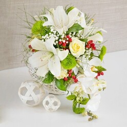 A luxurious white and green floral centerpiece is displayed as if ready to grace a wedding table in one of Oxford's historic venues, from Jericho townhouses to grand halls near the Bodleian. The flowers are gathered into a low, round white wicker basket that sits on a pristine white tabletop, framed by a softly textured beige background that feels warm and understated. Dominating the arrangement are several large white lilies, their generous trumpet-shaped blooms fully open, exposing fine speckling and golden anthers that hint at a gentle, sweet fragrance. Just above them, a single cream-to-pale-yellow rose holds a central position, its petals spiralling inward with quiet elegance. Flowing from the right-hand side, stems of white phalaenopsis orchids cascade down in a graceful arc, giving the design a waterfall or teardrop silhouette often chosen for wedding flowers. Around these focal blooms, vivid green pom-pom chrysanthemums create small, rounded bursts of texture, while feathery greenery and broader glossy leaves weave through the composition, adding depth and movement. Sprays of delicate white baby's breath drift between the larger flowers, softening transitions and lending a cloud-like lightness. Here and there, clusters of rich ruby-red berries stand out like tiny jewels, adding warmth and a touch of seasonal charm. On the left side of the image, two decorative white woven orbs rest on the table, echoing the basket's lattice pattern and enhancing the modern yet natural feel. The arrangement radiates purity, celebration, and thoughtful craftsmanship, ideal for same-day delivery across Oxford to bring refined beauty to a wedding breakfast, anniversary dinner, or special family occasion.