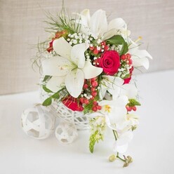 An elegant floral centerpiece, composed with the poise of a well-planned Oxford wedding, is displayed on a smooth white surface against a gentle, light beige fabric backdrop. At the heart of the design is a round, white, basket-style container, its open, lattice-like weave adding a handcrafted, almost lacework quality that feels right at home in a North Oxford townhouse dining room. From this airy base, tall white oriental lilies rise and unfurl, their broad petals creamy and luminous, each bloom showcasing proud stamens dusted with soft pollen. Between the lilies, deeply colored red roses rest like jewels, their velvety petals spiraling inward, bringing depth and warmth to the arrangement. Fine sprays of white baby's breath weave delicately through the larger flowers, creating a light, cloud-like veil, while slender, feathery greenery and bright green leaves lend freshness and soft movement. Scattered among the blossoms are shiny red hypericum berries, clustered like tiny lanterns that add a festive, seasonal feeling, ideal for a winter wedding reception at an Oxford college or a sophisticated celebration near Christ Church Meadow. Extending from the right side of the display, a cascade of white phalaenopsis orchids drapes gracefully downward, their rounded petals almost porcelain in appearance, with centers touched by faint yellow and blush tones, and several buds poised to open. On the left of the main design, three white woven spheres, each slightly different in size, rest on the tabletop and echo the texture of the container, visually tying the scene together. The overall impression is one of quiet luxury and romance, a centerpiece that would feel perfectly at home on a wedding table in Summertown or a special anniversary dinner near Oxford Castle.