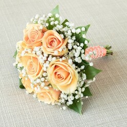 Bathed in the soft Oxford daylight, a lovingly gathered bouquet rests gracefully atop a lightly textured beige fabric, recalling the subtle tones of the city's ancient university buildings. At its heart, peach and apricot-hued roses-full bloomed with delicately spiraled centres-offer a tactile sense of layered elegance, their silken petals almost glowing against a lacework of dainty white gypsophila. These ethereal clusters create an airy balance, much like the tranquil Oxford Botanic Garden on a quiet morning. Glossy, deep green rose leaves peek with intention, grounding the pastel florals with a touch of natural vitality. The bouquet's round silhouette is completed with a neat coral-peach satin ribbon, which is artistically adorned by a trail of small, creamy white pearls twisting downward in a gentle swirl-suggesting an heirloom detail cherished through generations. The visible stem ends add to the rustic allure, reminiscent of bouquets curated for romantic strolls through Radcliffe Square. Flooded with even, soft illumination, the arrangement feels both substantial and whisper-light, perfect for a wedding aisle at the Sheldonian or an anniversary celebration overlooking the river Thames. This is an Oxford bouquet that lingers in memory-embodying the quiet sophistication, heartwarming joy, and enduring love that define cherished life moments within this historic city.