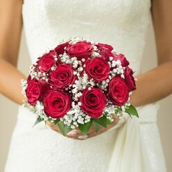 In this evocative moment on the edge of Oxford's dreaming spires, a bride's hands delicately cradle a domed bouquet of sumptuous, deep red roses, each rose unfolding its petals like verses in a love letter. The artistry is evident: every rose is nestled intimately alongside sprigs of dainty, snowy gypsophila, creating the impression of little constellations twinkling amid the crimson hues. Glossy green leaves provide grounding structure and a refreshing brightness, reminiscent of leaves glimpsed along the paths of Christ Church Meadow. The bouquet's passion meets pure grace as it rests against the bride's immaculate gown, which shimmers ever so slightly with the intricate patterns of lace or embroidery-its texture testament to the rich heritage of celebrations across Oxford. A soft ivory drape falls to one side, harmonizing effortlessly with the natural beauty of the florals. The scene is set in gentle, diffused light, the background fading to warm, neutral tones, evoking a hush of anticipation, like the first moments before saying I do in a centuries-old college chapel. This bouquet is more than floral artistry; it's the essence of Oxford romance-ideal for brides seeking a classic statement, or for articles on local wedding inspiration, floral design, or meaningful Oxford ceremonies.
