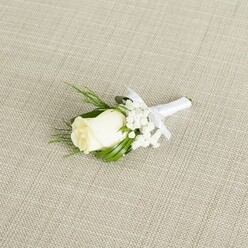 This image captures a classic wedding boutonniere resting on a pale, linen-like fabric that has a fine, crosshatched weave, similar to the soft table runners often used at reception venues around Jericho in Oxford. The boutonniere is built around a single creamy white rosebud, plump and perfectly formed, its petals smooth and gently curled inward as though protecting the promise of a full bloom. Subtle hints of green can be seen at the base where the sepals cradle the flower, adding a natural transition from stem to blossom. Surrounding the rose are airy clusters of white baby's breath, each tiny star-shaped flower creating a light, clouded border that softens the overall silhouette. A few elegant, narrow leaves in rich, glossy green and several more feathery sprigs of foliage tuck around and behind the main bloom, giving depth and a touch of fresh, garden-like movement, as if the flowers had just been gathered from a well-kept Oxfordshire garden. The stems of the rose and greenery are tightly wrapped with a bright white satin ribbon, smooth and slightly reflective under the gentle light, and finished with a neat bow tied just below the head of the rose. The diagonal placement across the neutral fabric background draws the eye along the length of the boutonniere, making it easy to imagine it pinned to a suit at a ceremony in one of Oxford's colleges or a registry office near St Aldate's. The mood is one of refined simplicity, conveying purity, new beginnings, and a quiet sense of occasion suitable for grooms, groomsmen, or any guest seeking a subtle yet meaningful floral accent.