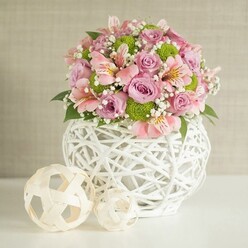 As I picture this stunning bouquet coming together on a quiet morning in Oxford, I'm struck by the interplay of textures and tones-a true study in touch and tranquility. The substantial, rounded arrangement rests gracefully upon a spotless white surface, aglow beneath soft, diffused light that feels like the gentle sun spilling through North Parade windows. Blooms of pastel pink and lavender roses speak of luxury and gentle affection, their lush, velvety petals arranged with expert care. These radiant roses are thoughtfully interspersed with pink alstroemeria, their trumpet-like flowers bearing subtle striped markings-each petal a miniature artwork reminiscent of Oxford's famed Botanic Garden in full bloom. Vivid lime-green button chrysanthemums spark playfulness and provide delightful contrast, their tightly clustered forms nestling amongst loose tufts of baby's breath, whose tiny white flowers deliver a dreamy, ethereal quality. Glossy, deep green leaves wind beneath and amid the flowers, anchoring the pastel palette with richness and depth. The contemporary white vase, created from intertwined strips in a delicate open weave, offers rustic charm with a modern sensibility-its tactile shape is echoed by two decorative rattan balls to one side, inviting both eye and hand to explore their textures. This centerpiece feels carefully curated for a celebratory table near Carfax Tower or a thoughtful gift for an anniversary celebrated along the banks of the Cherwell. Every detail, from the elegant lines to the fresh, abundant blooms, suggests quiet optimism and the joy of sharing beauty-and all crafted with the care and expertise of a true Oxford florist.
