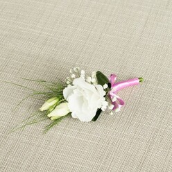 An elegant wedding boutonnière is photographed from a gently raised angle, resting diagonally across a light beige, linen-like fabric that brings to mind the textured tablecloths of intimate Oxford receptions. Dominating the composition is a single white eustoma flower, fully open and luxuriously ruffled, its velvety petals unfolding in soft circles that move from creamy white at the centre to a crisp, clean edge. Just to the left, two slender lisianthus buds in a pale green-cream hue nestle close, their pointed forms still tightly wrapped, suggesting the quiet promise of a ceremony about to begin in one of Oxford's historic churches or registry venues near the High Street. Scattered delicately around the main bloom are tiny sprays of gypsophila, or baby's breath, their numerous tiny white florets floating like mist and adding a light, romantic fullness. Behind the flowers, narrow blades of dark green, grass-like foliage arc outwards, drawing the eye towards the lower left corner and echoing the natural movement of stems in a garden just after rain. A single broader, deeper green leaf sits behind the central blossom, anchoring the design and giving the white petals a richer frame. At the bottom right, the stems are gathered and wrapped tightly with a striking fuchsia satin ribbon, tied in a neat, jaunty bow that introduces a bold splash of colour, reminiscent of a bright accent against a classic morning suit on an Oxford groom. The lighting is soft and even, revealing every petal curl and ribbon sheen, making this boutonnière feel freshly prepared for same-day wedding delivery across Oxford, from Summertown to Cowley, as a refined finishing touch to a carefully planned day.