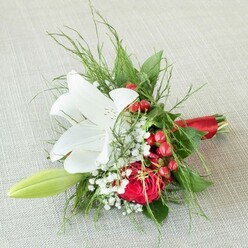 This harmonious bridal bouquet, artfully composed by an experienced local Oxford florist, lies elegantly against a natural, softly woven beige linen-evoking the textures found in Oxford's traditional shops near Cornmarket Street. At its luminous core, a single white lily fully blooms, its stately petals broad and silken with maroon speckling toward the center, drawing attention to vivid golden anthers. Nestled beside it, a deeply hued red rose radiates warmth, its plush petals whorled in a perfect spiral, stirring the heart like sunset hues reflected on the Isis. Scattered throughout are delicate clusters of white baby's breath, creating a dreamy softness, while vivid red berries offer a playful, festive punctuation reminiscent of seasonal décor in Jericho cafes and window displays. A slender, pale green lily bud gracefully extends to one side, symbolizing promise and new beginnings. The entire arrangement is encircled in a medley of textured greens-cascading, fern-like fronds and rich, sturdy leaves-giving a lush, garden-fresh impression that recalls Christ Church's blooming spring borders. Bound with a luxurious red satin ribbon, the gathered stems achieve a look both polished and heartfelt, perfect for bridal parties celebrating love in Oxford's storied surroundings. Bathed in gentle, even daylight, the composition's textures and hues are softly amplified, capturing both festive joy and gentle romance. This bouquet speaks quietly of cherished occasions and meaningful connections, designed to honour the beauty of weddings, anniversaries, or special moments throughout the Oxford community.