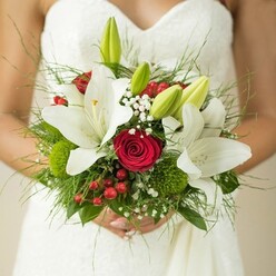 A bride's hands tenderly hold a captivating bouquet that blends classic sophistication with a dash of Oxford's garden charm. In a halo of soft, natural light, pristine white oriental lilies stand out, their elegant petals radiating innocence like the dreaming spires beyond. Deep red roses are gathered tightly at the heart of the bouquet, their lush, velvety spirals suffused with whispers of passion-a romantic tribute to timeless vows and riverside strolls through University Parks. Around them, clusters of tiny white Gypsophila lend a misty, ethereal texture, while unopened green lily buds evoke new beginnings, just as fresh as a dawn walk along Christchurch Meadow. Cheerful red hypericum berries and quirky green button-like blooms dance amid the feathery ferns and broad leaves, adding life and playful Oxford vibrance. The entire display is grounded by lush foliage, its deep greens mirroring the tranquil courtyards nearby. Every detail and contour is lovingly crafted, offering an arrangement that is both poetic and grounded in local tradition. This bouquet, arranged for a memorable Oxford wedding morning, promises to infuse the celebrations from Summertown to Headington with fragrance, colour, and hope-the perfect floral emblem from a local florist devoted to heartfelt moments.
