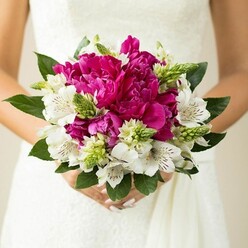 This close-up image focuses on a bride holding a compact, round wedding bouquet, perfectly framed against the soft white fabric of her gown, much like you might see outside Oxford's registry office or in the cloisters of a college chapel. At the centre, sumptuous peonies in vivid fuchsia and magenta create a bold focal point, their many ruffled petals layered so densely that they form a plush, cloud-like dome of colour. Nestled between these larger blooms, white alstroemeria (Peruvian lilies) add brightness and definition, each petal marked with fine maroon speckles and subtle striping that gives the arrangement a refined, almost painterly detail. Threaded through the bouquet are clusters of fresh green buds and spiky textural elements, perhaps early ornamental buds or filler flowers, which introduce a lively contrast in both shape and tone, evoking the feel of a walk through the greenery of South Park in late spring. Surrounding everything, generous deep green leaves cup the flowers and help create the bouquet's clean, rounded silhouette, their glossy surfaces catching the light. The bride's hands are seen just below the blooms, fingers gently wrapped around the stems, with nails neatly manicured in a pale blush shade that harmonises with the pink peonies. The background is softly out of focus, so the viewer's eye remains on the interplay of fuchsia, white, and rich green. The overall impression is one of joyful romance and polished craftsmanship, a bouquet that balances classic bridal elegance with vibrant personality - the sort of design brides across Oxford might choose for a summer celebration or a chic city-centre wedding.