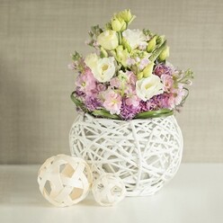 On a pristine white tabletop, a contemporary spherical vase cradles a lush spring bouquet, creating a tranquil focal point that would feel perfectly at home in an Oxford townhouse near Christ Church Meadow. The vase is crafted from slender white branches or painted rattan, woven into an open, basket-like globe that looks both light and architectural. Nestled inside, the flowers seem to billow gently, dominated by creamy white and pale lemon eustoma, or lisianthus, their velvety, multi-layered petals curling softly at the edges. Interspersed among them, stems of delicate pink stock rise in graceful spires, adding height and a subtle, sweet fragrance you can almost imagine drifting across the room. Below, clusters of vivid purple filler flowers form a dense, textural cushion that contrasts beautifully with the airy pastels above, while a neat ring of glossy green foliage frames the entire composition, giving it definition and a fresh, garden-picked feel. To the left of the vase sit two decorative spheres made from pale, natural rattan, their open lattice design mirroring the container and reinforcing the crafted, organic aesthetic. The background is a warm beige or greige wall with a fine vertical texture, perhaps fabric or wallpaper, which softens the scene and allows the gentle colours to stand out without distraction. Soft, diffused light falls across the arrangement, creating delicate shadows and a quiet, contemplative atmosphere, as if this centrepiece were waiting to grace a relaxed brunch table, a small wedding breakfast, or a thoughtful same-day flower delivery from Flowers Oxford to a loved one in Summertown.