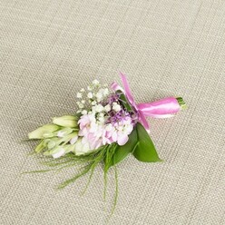 A single, carefully designed boutonniere or corsage is laid out on a finely woven, light beige linen backdrop, its subtle texture running diagonally like gentle ripples under the flowers. The focus is a nest of soft pink blooms, densely petalled and slightly ruffled, echoing miniature stock or garden-style flowers, their petals overlapping in delicate layers that look almost velvety to the touch. Threaded through this central cluster, delicate sprigs of white baby's breath form a mist of tiny blossoms, adding a cloud-like softness and giving the small arrangement a sense of space and airiness. Here and there, miniature spikes of lavender or deep purple florets emerge, adding depth and a more romantic, evening tone to the otherwise pastel palette, like the shift in sky colour over Christ Church Meadow at dusk. On one side, several elongated buds in creamy white fading to pale green are poised, still closed, suggesting freesias that will unfurl gently over the course of a celebration. The entire floral cluster is supported by glossy, rich green leaves that splay gently at the edges, while wispier threads of fine greenery curve outward, lending a natural, slightly whimsical feel. The stems are slim but tidy, bound together with a bold fuchsia pink satin ribbon wound smoothly around them, finished in an elegant bow whose loops and short tails glimmer softly under the light. This small but expressive piece would sit beautifully on a lapel at an Oxford wedding or graduation, a handcrafted detail one might order from Flowers Oxford for a ceremony near the Bodleian or along St Giles, quietly radiating joy and refinement.