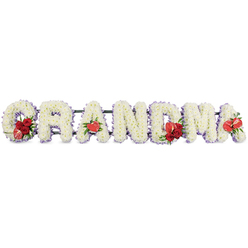 The image shows an elegant funeral flower arrangement spelling out "GRANDMA" in large, clearly defined block letters, the kind of personalised tribute often chosen by families across Oxford to honour a beloved nan or gran. Each letter is built entirely from dense clusters of pure white cushion chrysanthemums, their many-petalled heads packed closely together to create a soft, plush surface that feels both gentle and reassuring. Around the outline of every letter, a narrow edging of pale lilac or soft purple ribbon creates a subtle border, giving the tribute a calm, serene glow without overpowering the simplicity of the white blooms. Accents of vibrant red flowers are placed with great care to punctuate the design: on the "G", a lush cluster of deep red roses is nestled at one side, accompanied by a unique variegated red-and-cream calla lily that arcs gracefully over the white flowers. The first "A" carries a single heart-shaped red anthurium positioned low on the letter, its waxy petals and prominent spadix bringing a contemporary note. At the centre, the "M" is adorned with another grouping of rich red roses, balanced with an additional red anthurium to mirror the earlier motif. Towards the end, the final "A" is finished with its own red anthurium, visually linking the start and finish of the word. All the letters are mounted on a dark green metal or foam frame, which lifts them slightly and provides stability while remaining mostly hidden. Set against a plain white background, the tribute radiates love, respect, and quiet gratitude, perfectly suited for a memorial service or cremation farewell in and around central Oxford, Headington, or Cowley.