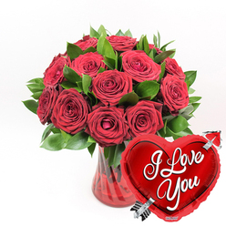 A luxurious romantic arrangement features a clear, fluted ruby-red glass vase brimming with deep red roses, gathered so closely together that they form a rich, velvety dome of colour. Each rose varies slightly in its stage of bloom: some are open and full, with satiny petals sweeping outwards, while others remain gently cupped, holding their fragrance and promise a little closer. Between the blooms, dark emerald foliage arches and curves, giving the design texture and contrast, much like the leafy avenues around University Parks in Oxford. The red glass vase itself has soft vertical ridges that catch and bend the light, lending a sense of movement and depth to the base of the display and subtly echoing the stems nestled within. Against a crisp white background, every shade of red and green appears especially vivid, as if the bouquet is illuminated by fresh morning light. In the lower right foreground, overlapping the line of the vase, a shiny, metallic red heart-shaped balloon leans in toward the flowers, as though drawn by their warmth. On its reflective surface, the words I Love You are written in flowing white script, easy to read and full of feeling. A stylised silver Cupid's arrow runs diagonally through the heart, symbolising love that has struck true. The image evokes the moment just before a surprise delivery arrives at a front door in Summertown or Headington: a classic pairing of roses and heart motif, ideal for marking anniversaries, Valentine's Day, or any occasion where love needs to be seen and felt immediately.