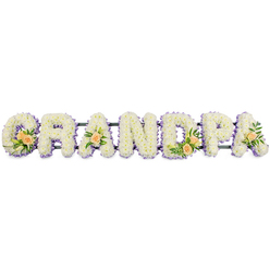 The image presents a beautifully composed funeral flower tribute spelling "GRANDPA" in bold, three-dimensional letters that stretch horizontally across the frame. Each letter is densely cushioned with small white chrysanthemums, their petals forming a uniform, velvety texture that looks almost like freshly fallen snow. Tracing the outer edges of the G, R, A, N, D, P, and final A is a fine outline of light lavender flowers, adding a soft lilac haze that gently separates the letters from the white background and lends a quiet, spiritual tone often appreciated for thoughtful services in Oxford's historic churches. At four points along the word, clusters of accent blooms bring warmth and individuality: the G and D each carry a small spray of peach roses, accompanied by slim white lily buds and sprigs of rich green foliage, while the first and last A are each adorned with a single peach rose and greenery. These touches of soft peach feel intimate and personal, like a private memory woven into the tribute. The entire word sits on a narrow, dark support bar, likely metal, which holds the letters in perfect line without distracting from the flowers themselves. Against the stark white backdrop, the tribute appears crisp, calm, and composed, ideal for resting at the front of a chapel or beside a photo at a memorial gathering in and around Oxford. The design balances formality with tenderness, expressing respect, remembrance, and enduring love for a grandfather who meant the world to his family.