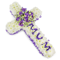 This substantial Christian cross is hand-constructed from thousands of compact white chrysanthemum florets, their rounded faces pressed closely together to form a plush, cloud-like surface that reads as reverent and pure against a stark white backdrop. A ruffled band of deep purple satin traces the outer edge, offering a refined border whose sheen contrasts gently with the matte chrysanthemum petals. The vertical arm bears the personalised message MUM, spelled out in small, concentrated purple blooms - likely lisianthus/eustoma or tiny spray chrysanthemums - the letters sculpted so they stand legible and intimate above the white field. The eye is drawn to a generous central spray at the cross's heart: tightly arranged violet and lavender roses, interleaved with soft purple lisianthus, sprigs of variegated greenery and small green berries that introduce depth and a natural, garden-like texture. The foliage shows lighter margins and deeper green centres, which catch the light and add crisp definition to the cluster. The overall mood is one of solemn beauty - cool hues that suggest peace, dignity and enduring affection. Shot in soft, neutral lighting, the tribute's textures and colour transitions are clear and calming. Prepared with local care in Oxford, this design is appropriate for memorials at St Aldate's or services near Jericho, and Flowers Oxford can arrange respectful delivery across the city when families request a piece that reads both personal and timeless.