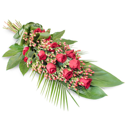 A refined, diagonal funeral spray is shown resting on a stark white background, its colours and textures carefully composed to express love and remembrance with quiet strength. At the upper left, a tight bundle of stems is wrapped smoothly in a broad green leaf, forming a polished handle that suggests the flowers have been freshly gathered by hand in an Oxford florist workshop before being placed at a service. From this point, the spray widens and then gently tapers, creating a long, flowing form that feels both structured and organic, reminiscent of a gentle river current moving through the city's peaceful parks. A line of rich, velvety red roses forms the focal path, each bloom different from the next: some are still modest buds, others partially unfurling, and several are fully open, with soft, layered petals that seem almost touchable. Threaded between the roses are clusters of small Hypericum berries, in warm coral-pink and red tones, their rounded shapes catching the light and adding a subtle shimmer. Deep green foliage cradles the flowers: broad, glossy leaves provide a strong, sophisticated background, while delicate palm fronds and finer greenery stretch outward, adding airiness and movement. The foliage varies in shade from dark forest green to brighter, fresher tones, hinting at renewal amidst grief. The entire composition feels respectful and composed, a traditional sympathy tribute suitable for a funeral in Oxford's churches, crematorium chapels, or quiet cemeteries, conveying sincere condolences and lasting affection in a single, graceful floral gesture.