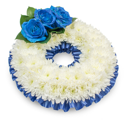Captured against a pristine white surface with a soft shadow beneath, this circular sympathy wreath is composed of a densely packed band of immaculate white chrysanthemums whose countless delicate petals form a gently domed, textured surface. From above, the full ring reads as an uninterrupted circle of peace and purity; the chrysanthemums' centers show the faintest hint of creamy yellow, adding a warmth beneath the cool whites. Artfully placed at the upper-left, a trio of deep royal blue roses creates a solemn, trustworthy focal point-each rose unfurling in perfect spirals, its velvet petals richly saturated and set among deep green, rounded foliage that cushions the cluster. A ruffled satin ribbon in matching cobalt trims the outer and inner edges of the wreath, its pleats catching light to introduce a subtle luster that frames the composition with dignified formality. The overall impression is of careful craftsmanship: measured spacing, balanced colour contrast, and thoughtful material choices that read as respectful and consoling. The bouquet's quiet perfume would be soft and reassuring rather than sweet, and the combination of textures-feathery chrysanthemum, velvet rose, glossy leaf, and silky ribbon-invites a contemplative response. Prepared for memorials across Oxfordshire, this piece is the sort of tribute we often assemble for services near Christchurch Meadow or deliveries to chapels in Jericho; Flowers Oxford can prepare and deliver similar wreaths with sensitivity and same-day consideration when needed.