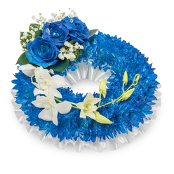 This funeral wreath presents a carefully considered study in blue and white: a full, circular field of tiny, royal-blue chrysanthemums packed so densely they create a tactile, cushion-like band of colour, each spidery petal contributing to a richly textured whole. A neat pleated white satin ribbon traces the inner edge, its sheen and soft folds offering an elegant, quiet contrast to the saturated blue. Crowning the upper-left curve are three commanding blue roses, their petals unfolding with a velvety thickness and deep sapphire tone; these are framed by delicate sprays of baby's breath and small, glossy dark green leaves, which add a classic, understated softness. Flowing from the mid-left across to the lower right, a graceful spray of white orchids - likely Phalaenopsis - arches gently, their smooth, pure petals and slender stems creating an airy counterpoint to the dense chrysanthemum base. Interspersed among the orchids are pale green buds at various stages, suggesting renewal and the passage of seasons. The composition is lit in soft, diffuse light that emphasizes texture over shadow, making the blues feel calm rather than stark. This wreath reads as a respectful, contemplative tribute, suitable for services across Oxford, from the peaceful lanes around University Parks to small gatherings near Jericho and Summertown. Prepared with careful craftsmanship by local hands, Flowers Oxford places value on balance, scent, and subtle movement when designing remembrances like this.