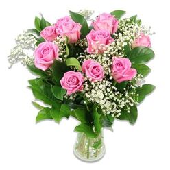 A classic glass vase overflowing with ten to twelve pink roses is shown against a bright, uncluttered white background, making every bloom and leaf stand out with crisp clarity. The roses themselves display a graceful range of pink tones, from pale blush at the outer petals to a slightly deeper, more saturated pink nestled at their centres, each flower layered like soft silk. Some roses are fully open, revealing their spiralled hearts, while others are in fresher bud stages, giving the bouquet a sense of life and gentle movement. Between the roses, airy sprays of white baby's breath float like tiny clouds, their delicate, snowflake-sized flowers adding texture and a light, celebratory feel that suits baby congratulations perfectly. Surrounding the blooms, rich green foliage frames the arrangement, the leaves slightly serrated and glossy, catching the light and anchoring the bouquet in nature. All of this is gathered in a clear glass vase, the stems neatly aligned, visible through the water as straight, vibrant green lines. The light in the image is soft yet bright, similar to daylight streaming through a window overlooking Oxford's peaceful residential streets, giving the flowers a freshly-picked appearance. The bouquet's rounded shape and balanced proportions suggest the hand of an experienced florist, thoughtfully composing each stem to evoke tenderness and joy. It is easy to imagine this arrangement from a trusted Oxford flower shop arriving at a maternity ward near the John Radcliffe Hospital, or being placed on a nursery windowsill in Cowley or Botley, its pink roses and white accents quietly celebrating a new baby girl and the love gathering around her.