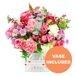 A vibrant mixed bouquet fills a clear, square glass vase, photographed against a spotless white backdrop so every detail stands out, as if displayed in a light-filled Oxford florist's studio. At its heart are generous pink roses, some a rich magenta with darker-tipped petals, others a softer, romantic blush that feels like a gentle embrace. These are nestled among plush, ruffled carnations in coral, peach-pink, and soft rose tones, their layered petals giving the arrangement a luxurious fullness. Dancing between the larger blooms are clusters of daisy-like chrysanthemums in shades of lavender and pale purple, each framed by luminous green centres that bring a lively sparkle, while creamy white flowers offer calm moments of light throughout the bouquet. The flowers are supported by abundant greenery: rounded eucalyptus leaves with their cool, silvery-green sheen, deeper green foliage adding depth, and a thread of reddish-brown leaves that hints at cosy autumn evenings by the colleges. The bouquet is presented in a transparent, rectilinear vase whose base is wrapped in a neat white band patterned with bright red hearts and the affectionate script all my love, making the intention of the gift unmistakable. In the bottom right corner of the image, an orange circular overlay clearly reads VASE INCLUDED in bold white text, reassuring anyone ordering same-day flower delivery in Oxford that this is a complete, ready-to-display present for birthdays, anniversaries, or simply brightening a home near Summertown, Headington, or along the Cowley Road. The overall effect is one of thoughtfulness, colour, and tender emotion.