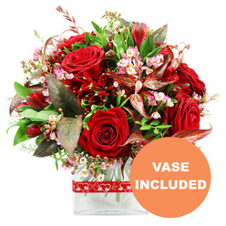 Against a pure white background, this romantic bouquet feels like a heartfelt love letter expressed in flowers, the kind of design a local Oxford florist might prepare for a proposal by the Radcliffe Camera or an anniversary dinner on Cowley Road. At the centre are sumptuous red roses, some tightly furled, others beginning to open into layered rosettes, their velvety petals creating soft shadows and depth. Nestled among them are smaller deep red blooms resembling alstroemeria or similar multi-petalled flowers, each one adding a sculptural quality and enhancing the passionate red palette. Threaded between these larger blooms are delicate sprays of tiny pale pink and white waxflower-style blossoms, their starry heads and slender stems lending a light, almost lacy texture. Around the flowers, an abundance of green foliage forms a generous, rounded dome: glossy leaves, finer sprigs, and arching stems all working together to frame the roses. Striking burgundy and reddish-brown leaves with gentle variegation peek through the greenery, introducing depth and a refined, dramatic edge that feels almost like the contrast of old stone and fresh greenery in Oxford's college gardens. The bouquet is anchored in a simple, contemporary clear glass vase with a rectangular base, allowing neatly cut stems to be seen and lending a polished, modern finish. A festive red ribbon printed with small white heart outlines wraps around the vase, adding a playful, clearly romantic touch. In the lower right corner, an orange circular label with bold white uppercase text clearly states VASE INCLUDED, signalling that this arrangement arrives beautifully presented and ready to display, perfect for same-day romantic flower delivery across Oxford.