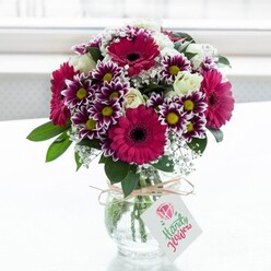 A lush, professionally arranged bouquet of mixed flowers is showcased in a squat, clear glass vase with a softly textured surface, placed against a bright, diffused white backdrop that suggests a sunlit Oxford window ledge. At the heart of the arrangement, large gerbera daisies in intense fuchsia and magenta glow like jewels, each dark, inky centre framed by layers of velvety petals. Around them, a ring of bicolour chrysanthemums blends deep purple edges into snowy white interiors, their fringed petals encircling warm yellow centres that add a joyful, almost playful note. Nestled between these bold blooms, creamy white roses and spray roses appear in different stages of opening, from tight buds to gently unfurling blossoms, adding softness, elegance, and the suggestion of a light, classic rose scent. Wisps of white baby's breath thread through the bouquet, airy and cloud-like, giving the whole design a sense of movement and breathing space above the glossy dark green foliage that anchors the composition. The stems are visible through the glass, neatly cut and fanned out, and the vase is tied with a rustic length of natural twine, knotted into a simple bow. A small, white rectangular gift tag hangs from the twine, decorated with a delicate pink rose symbol and flowing script, hinting at a personal note intended for the recipient. The bright, even lighting makes every colour and texture stand out, as though this bouquet has just been prepared by a local Oxford florist for same-day delivery to a home near Jericho, Summertown, or along Iffley Road, perfect for a birthday surprise, exam celebration, or a thoughtful pick-me-up. The overall mood is joyful yet refined, a bouquet that radiates warmth and care.