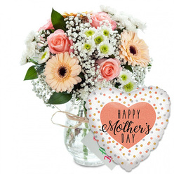 This Mother's Day ensemble is captured against a spotless white backdrop, where every blossom and reflective surface feels calm and intentional, like a still moment before family gathers for lunch in Summertown. The focal point is a lush, rounded bouquet resting in a clear glass vase, its sides smooth and simple, tied at the neck with a casual twist of pale twine that hints at countryside charm beyond Oxford's city streets. Within the bouquet, several peach-pink roses are fully open, their layered petals forming delicate spirals that invite you to lean in, while two or three generous gerbera daisies in warm peach-orange tones, each with a velvety dark brown centre, add a bold, sunlit presence. Between these stars of the arrangement, clusters of white baby's breath float like tiny clouds, and compact white chrysanthemums with subtle green hearts bring both softness and structure. Rich green foliage curves around the flowers, framing them as if they were gathered from a well-tended garden off Cowley Road. Nestled behind the blooms, almost hidden, sits a small white greeting card with a splash of green and pink handwriting, suggesting a heartfelt Mother's Day message from a child or partner. To the right of the vase, a large heart-shaped foil balloon leans forward with playful grace. The balloon's bright white surface is sprinkled with metallic gold and silver dots, mimicking scattered confetti, while a central peach heart carries the message HAPPY Mother's DAY in bold black letters, the word Mother's highlighted in flowing, calligraphic script. The combination of fresh flowers, thoughtful card, and celebratory balloon creates an image of warmth, gratitude, and gentle festivity, ideal for a same-day Mother's Day delivery across Oxford's neighbourhoods.