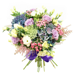 This opulent bouquet captures the heart of Oxford's garden charm-a lush tapestry of flowers gathered by hand near Magdalen College. At its core, ample hydrangeas in shifting shades of indigo and teal provide a cool, almost tranquil base. Tucked among them, lilac and lavender roses bloom with grace, their layered petals unfurling in soft spirals, evoking romantic strolls through Oxford's ancient lanes. Creamy white ranunculus and lisianthus peak from between the larger blooms, their intricate textures offering gentle firmness and depth, as if echoing the city's enduring scholarly legacy. Slender yellow lisianthus buds and chartreuse touches impart a fresh gleam, recalling delicate wildflowers in Christ Church Meadow. Wisps of vivid fuchsia and feathery pink astilbe lend a carefree spirit, trailing at the edges in a gentle breeze. Nestled throughout are sprays of fine, lacy green and white filler, reminiscent of Queen Anne's Lace, adding a touch of Oxfordshire's countryside to the composition. Framed by glossy dark green foliage, the bouquet gathers into a full, rounded form before the stems are elegantly tied with a royal purple satin ribbon-a nod to collegiate tradition. Suitable for a joyful celebration, heartfelt gesture, or as a sophisticated table centrepiece, this bouquet channels Oxford's poetic beauty and the attentive mastery of local florists, available for prompt delivery across the City Centre and Jericho.