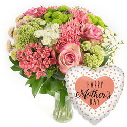 A lavish Mother's Day arrangement glows against a simple white backdrop, much like a bright shop window on Cornmarket Street catching the afternoon light. The flowers are gathered into a rounded bouquet and presented in a clear glass vase, the clean lines of the glass allowing the stems and clear water to show, hinting at the care taken by an experienced Oxford florist. At the heart of the design, several large pink roses unfurl, each petal gently curling and edged with a slightly darker hue, creating a soft, romantic focal point. Around them cluster small coral-pink flowers reminiscent of kalanchoe, tightly grouped to create pockets of texture and warmth. Lime-green button chrysanthemums stand out with their compact, neatly domed heads and fine, feathery petals, bringing a bright, modern twist to the otherwise classic palette. Interwoven through the bouquet are airy white spray chrysanthemums, their daisy-like faces adding lightness and a sense of space, while tiny spiky coral blooms and occasional chartreuse green florets tuck into the greenery, adding layers of interest when viewed up close. Rich, dark green foliage cradles the blossoms, framing the colours and giving the impression of a carefully curated garden in miniature, the kind you might discover in a quiet courtyard off St Giles'. To the lower right, a heart-shaped foil balloon completes the gift. Its glossy surface reflects the light, and its white or pale silver ground is dotted with shimmering gold, silver, and peach confetti-style spots. In the centre, a peach heart bears the message HAPPY Mother's DAY in black script and bold type. The overall mood is one of festive gratitude and affection, a ready-to-deliver Mother's Day surprise for homes across Oxford, from Summertown to Cowley.