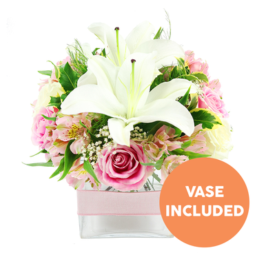 A sophisticated floral centerpiece is captured against a clean white background, its compact yet abundant design radiating a sense of calm elegance, like a well-kept townhouse garden off the Cowley Road. In the middle of the arrangement, two pristine white Oriental lilies dominate, their wide, star-shaped petals gently arching outwards and revealing tall, dark reddish-brown stamens that add an artistic contrast. Encircling the lilies is a generous collar of pastel and soft pink roses, some tightly furled in classic spirals, others just beginning to relax their petals, and a few fully open with ruffled edges, suggesting both new beginnings and lasting affection. Interwoven between the roses are sprays of pink and white alstroemeria, each bloom marked with fine, speckled strokes of yellow on its inner petals, lending a painterly, almost watercolour quality. Wisps of white baby's breath hover between the larger flowers, giving the design an airy, floating lightness, like blossom drifting along the River Cherwell in spring. A tapestry of green foliage-broad, shiny leaves and more delicate, feathery greens-surrounds and supports the blooms, adding texture and a sense of freshness you can almost smell. The flowers are expertly gathered into a low, square clear glass vase, whose straight edges contrast pleasingly with the rounded dome of the bouquet. Around the vase, a soft pink satin ribbon is tied horizontally, offering a subtle decorative detail that makes the design feel ready to place straight onto a dining table in Headington or a cosy living room in central Oxford. In the lower right corner of the image, a bold orange circular graphic with crisp white text clearly announces VASE INCLUDED, making it visually clear that the container comes with the arrangement. The entire composition feels serene and thoughtful, perfect for sending gentle congratulations, sympathy, or simply a message of appreciation with local flower delivery in Oxford.