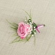 A single pink rose boutonniere lies on a softly textured light beige linen fabric, captured from a gently elevated angle that lets you see every petal and detail as if it were resting on a dressing table before a wedding at an Oxford college chapel. The rose is full and freshly opened, its velvety petals shifting from a warm blush at the centre to a slightly deeper pink at the curled outer edges, giving the impression of a romantic, just-cut bloom. Around its base, tiny clusters of white baby's breath form a delicate, cloud-like halo, their miniature blossoms adding lightness and a classic, almost vintage elegance. Wispy strands of fine, grass-like foliage in a fresh spring green fan out from behind the rose, creating a soft frame that draws the eye inward, while a single broader, darker green leaf is neatly folded to cradle the stems, lending structure and a polished, tailored look typical of a professional florist's hand. The stems are tightly bound with a smooth pink satin ribbon, tied into a small, tidy bow that echoes the rose's hue without overpowering it. A slim, silver-toned pin is visible near the base, signaling that this piece is ready to be fastened to a lapel for a groom, groomsman, or prom guest heading out from central Oxford or along the Cowley Road. Gentle, even lighting casts soft shadows across the natural linen, highlighting the boutonniere's three-dimensional shape and the interplay of textures-plush petals, airy gypsophila, sleek ribbon, and woven fabric-capturing a moment of quiet anticipation before a day of celebration and photographs.