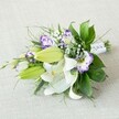 A hand-tied bouquet of white lilies and purple lisianthus lies diagonally across a softly textured light beige fabric, as if resting on a calm windowsill in an Oxford townhouse near the High Street. The focal point is a cluster of large Oriental lilies, their pure white petals opening generously to reveal yellow-orange stamens dusted with pollen and faint reddish speckles that draw the eye inward. Around them, several vivid chartreuse-green buds are still tightly closed, promising more blooms in the days to come and giving the arrangement a sense of gentle movement and growth. Threaded between the lilies, ruffled lisianthus flowers in white edged with lilac and deep violet create a painterly effect, like brushstrokes of purple against cream. Tiny clouds of white Gypsophila float between the larger blooms, softening the composition and adding an airy, bridal lightness. The flowers are framed by lush greenery: broad, glossy dark green leaves that provide structure, interlaced with finer, wispy fronds and a few spikier stems that lend texture and a slightly wild, garden-picked feel, reminiscent of walks through University Parks in late spring. At the narrower end of the bouquet, the stems are neatly gathered and wrapped in a crisp white satin ribbon, delicately accented with small, pearl-like beads that catch the light without overpowering the flowers. The soft, diffused lighting in the image enhances the creamy whites and cool purples, without harsh shadows, giving the bouquet an elegant, serene mood, ideal for a bridal party, a refined gift for an Oxford college celebration, or a graceful centerpiece for an intimate dinner near Jericho.