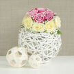 The image presents an elegant floral composition styled as a full-frame scene, with no distractions beyond a luminous white tabletop and a soft, neutral beige backdrop textured like fine linen. At its heart stands a lush, spherical bouquet of roses, carefully sculpted into a compact dome that feels both intimate and luxurious. The roses alternate between soft, romantic pink and buttery, creamy yellow, each blossom full and velvety, petals overlapping in generous spirals that suggest freshly cut stems from a trusted local florist. Wisps of delicate white baby's breath weave gently around the roses, creating a misty, cloud-like border that brings a sense of airiness and tenderness, like light fog over the Thames near Folly Bridge in Oxford. A few understated green leaves are tucked low at the base, just visible enough to remind the viewer of the living garden roots of this refined design. The bouquet is cradled in a distinctive, modern vase formed from interlaced white rattan-style strands. This open, spherical structure feels like a minimalist basket or a sculptural orb, its gaps allowing glimpses of shadow and light that dance across the white surface below. To the right, two smaller decorative orbs made from pale, natural wooden strips echo the same woven aesthetic. The larger of the pair rests slightly behind, while the smaller nestles closer to the foreground, creating depth and a gentle, rhythmic balance. Soft, even lighting enhances the pastel tones and smooth textures, lending an atmosphere of tranquil celebration. The arrangement would suit an Oxford townhouse near Summertown, a cosy anniversary dinner, or a refined welcome-home gesture, radiating thoughtful craftsmanship and understated luxury.