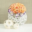 A romantic floral centerpiece is displayed on a glossy white surface, bathed in gentle, even light that could be streaming into a quiet Oxford dining room before guests arrive. The arrangement is dominated by a perfectly rounded dome of roses in tender shades of peach, apricot, and soft blush, packed so closely that their overlapping petals create a smooth, cushion-like form. Each rose appears at its peak, with spirals unfurling from the centre and outer petals slightly relaxed, suggesting freshness and subtle fragrance. Nestled around the base of this rose dome, delicate lisianthus blossoms unfold in creamy white, their frilled petals edged with vivid violet and purple tones. These bi-coloured edges deliver a refined contrast, while wisps of white baby's breath thread between the lisianthus heads, adding a fine, cloud-like texture. The flowers are set into a sculptural white container woven from thick strands of wicker or rattan, swelling into a rounded, bulbous shape. Its open lattice design allows glimpses of shadow and light to play through, echoing modern yet natural craftsmanship. In front of the vase rest two decorative orbs, one medium and one larger, made from pale, natural wood-toned strips interlaced into airy spheres that mirror the basket's texture. The entire scene is framed by a soft beige fabric backdrop, slightly textured like linen or canvas, which offers a warm, understated canvas for the pastel blooms. The overall mood is understated luxury and gentle celebration, ideal for envisioning wedding table arrangements, anniversary dinners, or intimate gatherings held across Oxford, from riverside restaurants to hidden courtyard venues.