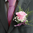 This image shows an elegant wedding boutonniere fastened to a charcoal or deep navy suit, the kind a groom or groomsman might wear to a ceremony in central Oxford or along the river near Folly Bridge. At its heart is a single pale pink rose, petals unfurling in a graceful spiral, each layer soft and velvety, as if brushed with morning light filtering through college cloisters. Surrounding the rose, delicate sprays of white gypsophila form a light halo, their tiny blossoms adding a feathery texture that softens the strong line of the lapel. Wisps of fine green foliage arc and weave around the composition, lending movement and a fresh, garden-like feel, as though the flowers have just been gathered from an Oxfordshire country garden. The stems are bound by a neatly tied ribbon in a muted lilac or mauve shade, the satin finish catching subtle highlights and providing a gentle link between the rose and the darker tones of the suit. The lighting is soft and even, allowing the blush of the rose to appear almost radiant while the greens and whites remain crisp and distinct. You can almost imagine the quiet rustle of fabric as the wearer adjusts their jacket before stepping into a ceremony at a hidden chapel off the High Street. Crafted with care and precision, this boutonniere expresses romance, composure, and gentle tradition, the kind of finishing touch a local florist in Oxford would create for an intimate wedding, civil ceremony, or special celebration shared with close family and friends.