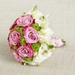 A luxurious, hand-tied bridal bouquet is photographed from a gently elevated angle, resting on a softly textured light beige fabric that feels like a calm Oxford morning. At the centre of the rounded arrangement, several large roses open fully in a romantic lavender-pink, their velvety petals deepening to richer fuchsia tones at the edges, creating a subtle two-tone effect that draws the eye. Threaded between these statement roses are clusters of bright chartreuse green button chrysanthemums, their compact, cushion-like heads adding a crisp texture and lively freshness that recalls new growth along the River Cherwell in spring. Delicate white blooms, likely lisianthus, are scattered throughout, their layered, ruffled petals and tiny greenish-yellow centres lending an airy softness and a whisper of classic bridal elegance. A few tight green buds peek through, hinting at just-cut freshness, as though the bouquet has come straight from an early visit to Oxford's Covered Market flower stalls. The stems are gathered neatly and wrapped in smooth pink satin ribbon, forming a comfortable handle for the bride to hold as she walks down the aisle at a city centre church or a college chapel. Spiralled over the ribbon is a fine strand of small white pearls, adding a vintage, heirloom touch that feels perfectly suited to Oxford's historic streets. The soft, diffused lighting enhances the pinks, whites, and greens, creating a bouquet that radiates romance, celebration, and refined floral craftsmanship-an ideal visual for modern wedding inspiration or elegant gifting within the Oxford area.