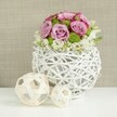 An artfully composed floral dome of pastel roses and fresh greenery is displayed in a white woven vase on a crisp white surface, lit with soft, even light that flatters every petal. The vase, spherical and slightly flattened at the base, is crafted from slender white twigs or rattan strands crisscrossed into an open lattice, lending a modern yet organic presence that would feel at home in a stylish North Oxford living room. Emerging from this airy base, fully opened roses in soft blush pink and muted lavender tones take centre stage, their petals lush and velvety with subtle hints of deeper pink tracing the edges. Interspersed among them, bright green button chrysanthemums offer small bursts of vivid colour and a finely textured surface, bringing energy and contrast to the arrangement. At the lower edges of the bouquet, small white and cream filler blossoms-resembling baby's breath or tiny spray roses-gently spill over, creating a halo effect that blends the flowers into the sculptural vase. To the left, two decorative spheres in light, natural wicker or wood sit on the same white tabletop, one larger, one smaller, both with an open, woven construction that mirrors the vase and adds a crafted, tactile detail. Behind, a softly blurred panel of beige or tan fabric, textured like linen, creates a warm, neutral backdrop reminiscent of wedding linens in an Oxfordshire country venue. The overall mood is refined and tranquil, an ideal floral centerpiece for a wedding reception table, engagement dinner, or elegant home gathering, conveying care, freshness, and thoughtful local design.