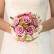 A radiant wedding bouquet is the centrepiece of this tender Oxford moment, cradled by a bride whose gown shimmers with intricate lace detail found in historic venues along the High Street or near the Sheldonian Theatre. The bouquet is a masterful interplay of colour and light: at its heart, plush roses bloom in delicate shades of dusty mauve and lavender pink, their petal edges kissed with vibrant fuchsia that catches the gentle glow of an English spring. Encircling the main blooms are round, lime-green button chrysanthemums, their compact, playful texture providing an enlivening contrast against the powder-soft pastel roses. The outer rim of the bouquet is expertly finished with tiny, fragrant white blossoms-sprays of rose or freesia-intermixed with hints of green, evoking the fresh promise of a new beginning. The bride's hands, polished in subtle pink with a joyful touch of shimmer, cradle the flowers with evident delight. Soft, even lighting enhances the tactile beauty of every element-lace, petal, and leaf-crafting an atmosphere of calm anticipation and romance. This bouquet, crafted for weddings and celebrations across Oxford and its charming boroughs, encapsulates the artistry, joy, and timeless grace of a very special day in the heart of the city.