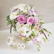 An artfully arranged floral display fills a round white wicker basket, captured in a luminous, close-up composition that feels ready to be placed at the centre of a wedding table in Oxford. The bouquet is dominated by lush pastel pink roses, each one fully open so their spiral hearts are clearly visible, the petals layered like silk and gently curling outward. Weaving through the roses are generous white Phalaenopsis orchids, their broad, sculptural petals and golden-yellow centres adding a refined, modern touch that contrasts beautifully with the classic roses. Ruffled white Lisianthus with faint lilac-tinged edges and softly scented pale pink stock blooms cluster between the larger flowers, some stems gently spilling over the basket's rim to give a sense of effortless movement. Sprays of delicate white baby's breath thread through the whole design, breaking up the fuller heads with tiny dots of light that catch the eye and soften the overall texture. Fresh green foliage and small buds emerge from between the blossoms, bringing a lively, botanical feel, as if the flowers were just arranged by hand in a local Oxford florist studio. The basket sits on a bright white surface, alongside two decorative woven spheres in pale, natural wood tones that mirror the wicker's texture and lend a calm, organic accent. Behind the arrangement, a softly textured, neutral beige background fades into blur, allowing the delicate colours and shapes of the flowers to stand out. The image conveys romance and quiet celebration, perfectly suited to illustrate wedding table arrangements, anniversary flowers, or elegant home decor for gatherings across Oxford.