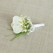 An elegant white orchid boutonniere rests quietly on a softly woven, light beige fabric, the texture reminiscent of natural linen often seen in historic Oxford townhouses. At the heart of the piece, a pristine Phalaenopsis orchid bloom opens with calm confidence, its broad, velvety petals a pure, cool white that seems to glow against the neutral background. In the centre of the flower, a gentle flush of pale, buttery yellow in the throat adds a hint of warmth, suggesting freshness and life. Clustered closely around the orchid, airy tufts of white baby's breath create a delicate, cloud-like halo, their tiny blossoms forming a fine, misty texture that softens the edges of the design. Interwoven among the gypsophila are slender strands of feathery green foliage, possibly asparagus or maidenhair-style fern, their bright, fresh green tones evoking the leafy calm of Oxford's University Parks after a spring shower. The stems of the orchid, baby's breath, and greenery are expertly gathered and wrapped in a smooth white satin ribbon, tied into a neat, modest bow at the base to keep the form compact and ready to pin onto a lapel. The lighting is gentle and even, without harsh shadows, allowing the whites and greens to appear clean and true to life. The overall mood is one of quiet refinement and timeless sophistication, the sort of classic boutonniere a groom might wear at an Oxford college chapel wedding or a formal ceremony near the Bodleian Library, bringing together purity, subtle texture, and understated style in a small, perfectly balanced floral accent.