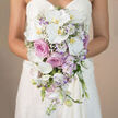 Framed at waist height against a softly blurred beige backdrop, a bride in an elegant white sweetheart-neckline dress holds a lavish waterfall bouquet, the flowers spilling gracefully from her hands in a long, tapering arc. The colour story is gentle yet luxurious, blending crisp white, warm pink, and muted lavender in a palette that would sit beautifully against the stone walls and leafy quads of Oxford's historic colleges. Dominating the design are broad, silky Phalaenopsis orchids, their white petals curving outward to reveal glowing yellow-orange centres that add a hint of tropical glamour. These sculptural blooms are interwoven with full-petalled pink roses and more textured, slightly ruffled lavender-pink garden roses, each rose capturing the eye like a small, gathered cloud of colour. Around them, clusters of white lisianthus with soft lilac edges and airy stems of white gypsophila create lightness, giving the bouquet an almost misty halo. Slender spires of pale lavender stock add height and rhythm, extending the cascade and suggesting a gentle movement, as if the flowers are swaying with each step along the River Thames at Folly Bridge. The overall impression is romantic and meticulously crafted, yet relaxed, as if the bouquet has grown naturally into its teardrop shape. Every element appears fresh and dewy, as though just arranged for same-day wedding delivery in Oxford, reflecting the care of a florist who knows how to balance opulence with restraint for ceremonies held in churches, registry offices, or intimate garden venues across the city.