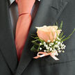 Pinned to the left lapel of a deep charcoal suit, a single peachy-pink rose becomes the centrepiece of a restrained, romantic boutonnière photographed in gentle, natural light. Each petal is rendered with tactile detail-soft, slightly ruffled edges and a subtle gradient from warm peach to paler inner folds-inviting a sense of touch as much as sight. Tiny clusters of white baby's breath float like miniature clouds around the rose, while slender, feathery sprigs of green foliage add a crisp, organic outline that lifts the bloom away from the structured fabric beneath. The arrangement is bound at its stem with a fine blush satin ribbon, its pale sheen mirroring the tie's warm hue and resting against a light dress shirt patterned with a faint dark grid that gives the ensemble quiet geometric interest. The overall mood is refined and personal, a floral whisper that balances ceremonial polish with handcrafted warmth. You can picture this detail on the groom at a Botley Road venue or on a best man stepping out from a rehearsal near Summertown, an emblem of delicate craftsmanship that carries the fragrance of garden roses and the soft attentiveness of a local florist preparing thoughtful, wearable flowers for special days in Oxford.