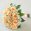 There is a timeless tenderness to this round hand-tied cluster of peach-hued roses, their creamy-apricot petals layered like soft silk and catching the pale, diffuse light. Clusters of tiny white gypsophila drift between the roses, adding an ethereal, cloud-like lift that balances the weight of each larger bloom. Oversized, glossy green leaves are arranged to cradle the composition, introducing a deep emerald contrast that grounds the pastel story and gives the bouquet an organic silhouette. The stems are gathered and wrapped in a textured peach ribbon scattered with pearls, a finishing touch that feels thoughtful, slightly vintage and perfect for gentle, ceremonial moments. Close your eyes and you'll sense the fragrance: a restrained rose perfume brightened by the clean, green notes of fresh foliage and the whisper of gyp, subtle enough not to overwhelm but present as a comforting memory. As an Oxford florist I picture this carried out of the Covered Market on a spring morning, or presented after a small ceremony near Radcliffe Camera - it suits weddings, vow renewals and understated anniversaries. The lighting in the photograph flattens any harsh shadow, allowing the soft colours and rich textures to speak clearly; each petal, each pearl-studded wrap reads as a deliberate, careful decision by the maker. Delivered locally across Oxford, this bouquet is both a visual and tactile keepsake, an object of quiet celebration that will age gracefully in memory as well as in vase.