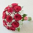 A striking bridal bouquet is presented against a soft, neutral background, its lush, deep red roses and delicate white baby's breath arranged in a splendid, harmonious round. The roses, double-layered and at the height of their bloom, radiate rich, velvety texture-a passionate centerpiece loved by many brides in Oxford. Sprinkled throughout, clusters of tiny, airy gypsophila provide clouds of brightness and softness, echoing the picturesque spring gardens in University Parks. Subtle glimpses of dark green foliage peek through, grounding the impression with nature's freshness. The hand-tied bouquet is finished with immaculate detail: a wide satin ribbon in brilliant red, carefully striped in white, and an elegant winding of luminous pearl beads. The freshly cut stems, visible at the base, hint at the skilled touch of an Oxford florist, their neatness inviting the bouquet to be cradled by the bride. Basked in natural daylight, every hue and intricate petal stands out, making this arrangement equally suitable for wedding days, anniversaries, or romantic celebrations-especially for those in Oxford seeking a floral gesture that resonates with both classic sentiment and the unique local spirit.