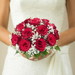 In this image, a bride holds a classic, hand-tied bouquet of red roses and white baby's breath, the arrangement glowing with timeless romance in an Oxford setting. The roses are a deep, sumptuous scarlet, each bloom at its prime, with velvety petals curving inward to form full, rounded heads. Interlaced between them, fine sprays of baby's breath create a gentle veil of white, their tiny star-shaped blossoms adding lightness and a feathery texture, like a dusting of early-morning frost. A collar of fresh green leaves encircles the base, giving the bouquet structure and a natural frame that hints at gardens tucked behind old stone walls or along the paths of the Botanic Garden.
The bouquet's silhouette is distinctly round and thoughtfully composed, revealing the steady hand of a skilled wedding florist. There is harmony in the way each rose sits snugly beside the next, no gap left unattended, no stem out of place. The rich crimson stands in poignant contrast against the bride's pale gown, perhaps lace-trimmed or satin-smooth, creating a striking visual echo of love and celebration. You can almost sense the weight of the bouquet in her hands, firm yet comforting, as she pauses for photographs on an Oxford street near Broad Street or by a college doorway. The subtle fragrance of roses and greenery seems to linger in the air, promising warmth, devotion, and the start of a shared life. The photograph captures not only a floral design, but a heartfelt symbol of promise on a wedding day shaped by the charm and history of Oxford.