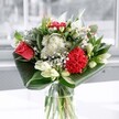 A contemporary mixed bouquet in a clear glass vase stands in soft window light, its colours and textures unfolding like a quiet celebration in an Oxford townhouse. At the heart of the design, a single coral-red rose and a plush crimson carnation glow with velvety depth, their layered petals suggesting warmth, devotion, and the thrill of good news shared over coffee on the High Street. Around them, clusters of delicate white chrysanthemums and daisy-like blooms form a gentle halo, each tiny petal catching the cool daylight and softening the intensity of the reds. Creamy white lilies, some fully open and others still in smooth, tapered buds, rise gracefully through the arrangement, their star-shaped flowers hinting at a light, sweet fragrance that feels as fresh as a walk along the River Thames near Folly Bridge. The bouquet is framed by deep emerald leaves, folded and arched by an experienced florist to cradle the flowers in sweeping curves, giving the piece a modern, sculpted silhouette while still feeling natural and inviting. The glass vase reveals clean stems and clear water, underscoring the sense of freshness and care that went into its creation. Reflections of the window play across the vase and glossy foliage, adding subtle highlights and depth. This is the kind of floral design you might send for a birthday, a new home in Jericho, or simply to brighten a friend's desk near Oxford city centre, a radiant, uplifting arrangement that brings texture, light, and a feeling of thoughtful generosity into any room.