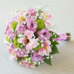 Laid gently on a pale, textured linen surface that looks like it could dress a refined Oxford dining table, this lush bouquet forms a rounded, almost domed shape, brimming with layered colour and texture. The central focus is a collection of lavender roses, each rose opening in generous spirals, the petals curling softly at the edges to create a velvety, almost luminous presence. Threaded between them are pink alstroemeria blooms, their narrow petals stretching out like tiny wings, marked with subtle streaks of rose and hints of lemony yellow, adding movement and detail that draw the eye. Tucked playfully among these are chartreuse green button chrysanthemums, their compact, spherical heads offering a bright, contemporary shock of colour, a vivid green that feels as fresh as the lawns around Christ Church Meadow after rain. Fine sprays of white baby's breath scatter delicately throughout the bouquet, their miniature blossoms catching the light and giving the arrangement an airy shimmer, as though a light mist has settled over it. The stems are tightly bound with a glossy, wide satin ribbon in a pink tone that echoes the flowers, and a neat wrap of tiny white pearls runs along the handle, providing a refined, bridal accent. The natural stems extend past the wrapping, bringing in an organic, hand-gathered feel, as if the bouquet has just been collected from a flower stall off the High Street. Crafted with care, this composition suits Oxford registry office weddings, college chapel ceremonies, or romantic proposals on the banks of the Cherwell, offering a sense of modern romance rooted in classic floral design.