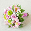 Viewed from above on a softly textured, neutral cloth that resembles natural linen, this bouquet appears as though it has just been set down between wedding photos and champagne flutes at an intimate Oxford celebration. The central focus is on full, pastel-pink roses, their petals unfolding in gentle spirals that suggest tenderness and quiet romance. Around them, round lime-green button chrysanthemums provide a refreshing jolt of colour, their intricate, cushion-like heads introducing a crisp texture that contrasts beautifully with the smooth rose petals. Pale pink alstroemeria weave in and out of the design, their inner petals etched with fine raspberry streaks and dappled with yellow, giving the bouquet a painterly, almost watercolour feel. Wisps of delicate white baby's breath are tucked into every gap, lending an airy, cloud-soft finish that keeps the arrangement light and ethereal rather than heavy or formal. Lush green foliage hugs the outer edge, curving gently outward as though cradling the blooms, evoking the leafy calm of University Parks or a shaded corner of Headington. The stems beneath are gathered neatly and wrapped firmly in a rich fuchsia satin ribbon, its sheen catching the soft light and suggesting care and craftsmanship. The overall mood is hopeful and celebratory, ideal for an Oxford bride, a proposal along the Isis, or a heartfelt delivery to a loved one in Summertown from a florist who understands both flowers and the rhythm of local life.