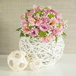 In this image, a lush, dome-shaped bouquet is presented as a modern centerpiece, as if ready to grace a sunlit Oxford living room overlooking Christ Church Meadow. The flowers are densely arranged yet carefully balanced, with full-bloom roses in soft lavender and rich pink tones forming the main body of the design. Their layered petals curl outward in generous spirals, giving the arrangement a sense of quiet opulence. Between them, you can see pale pink alstroemeria with their familiar speckled throats, each bloom adding a sense of graceful movement, like brushstrokes of colour dancing through the bouquet. Nestled closely around the roses are rounded clusters of vivid lime green button chrysanthemums, whose tightly packed, textured heads create a refreshing contrast to the smooth rose petals. Wisps of white baby's breath weave through the gaps, their tiny blossoms creating a delicate, misty veil that softens the bolder shapes. Deep green leaves are mostly tucked beneath the blooms, just visible enough to add depth and a natural frame. The flowers rise from a striking spherical vase crafted from white woven wicker or rattan, its open, airy lattice reminiscent of the intricate stonework of Oxford's colleges, but translated into a clean, contemporary form. This sculptural vase rests on a smooth, white surface, which reflects gentle highlights from the arrangement. Beside it sit two lighter, natural-toned woven balls, echoing the vase's pattern and adding a subtle, organic décor touch. The background is a calm, neutral wall, allowing every blossom and texture to stand out clearly, creating an atmosphere of thoughtful design, gentle joy, and refined simplicity, ideal for weddings or intimate gatherings across Oxford.
