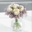 A close-up photograph captures a refined white and lavender bouquet resting on a smooth white surface, the entire arrangement bathed in soft natural light that feels very much like a calm morning in Oxford. At the heart of the design are several creamy white roses, their petals unfurling in flawless spirals, with just a whisper of pale buttery yellow deep in the centre that suggests they were cut fresh only hours ago. Around these roses swirls a cloud of tiny lavender-purple blooms, likely statice or limonium, their papery texture and clustered form creating a misty, romantic halo that softens the whole composition. Each stem is gathered neatly into a clear glass vase with a distinctive crackle, or ice-effect, pattern, so the surface seems to shimmer as it catches the light, throwing delicate highlights and reflections onto the bright tabletop below. Through the transparent water, you can clearly see the straight, green stems crossing and overlapping, grounding the bouquet and adding a sense of natural structure. The background melts into a gentle, blurred grey, reminiscent of an overcast sky above the Oxford spires, ensuring the eye remains focused entirely on the flowers. The mood is tranquil and contemplative, ideal for a quiet reading nook overlooking the gardens off Iffley Road, for a gentle thinking-of-you gift, or for bringing a sense of peaceful elegance to a small workspace. Every element, from the colour balance to the way the light glows on the rose petals, conveys understated grace and the steady hand of a florist who understands both texture and emotion.