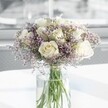 A generous, rounded bouquet of creamy white roses and soft lavender statice stands in the centre of the scene, poised in a tall, clear glass cylinder vase on a bright white tabletop. Around a dozen roses, perfectly formed and freshly opened, show layers of velvety petals in shades that hover between warm ivory and gentle cream, each bloom facing slightly differently so the bouquet feels natural rather than rigid. Between the roses, countless tiny star-shaped blossoms of pale lavender-purple statice create a filigree cloud, their papery texture and branching stems adding a delicate haze that softens the composition. The green stems, neatly cut and gathered, are visible through the vase, submerged in clean water that catches the light and creates faint reflections on the glass. Soft daylight filters in from an unseen window, much like the calm light in an Oxford townhouse near the High Street, bathing the flowers in a calm glow and casting the faintest shadows on the smooth surface beneath. The background is blurred and neutral, so all attention rests on the bouquet's soothing palette and elegant form. The arrangement feels serene and composed, ideal for a thoughtful sympathy gesture, a quiet anniversary surprise, or a peaceful centrepiece for a study overlooking Christ Church Meadow. The imagined scent is subtle and refined-a whisper of fresh rose blending with the green, slightly herbal note of the stems-suggesting both freshness and restraint. This image captures the kind of timeless, understated design a careful Oxford florist might prepare for same-day delivery to bring calm and comfort into a contemporary home or office.