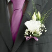 Pinned against the charcoal fabric of a tailored wedding suit, this boutonnière feels like a quiet breath of elegance at a ceremony in central Oxford. At its heart sits a single open lisianthus flower in pure white, its petals layered and slightly frilled, like folded silk or fine parchment pages in an old Bodleian library book. The bloom is full but never showy, conveying purity and calm confidence. Behind it, two upright lisianthus buds emerge, pale and slender, holding the promise of blossoms still to come and echoing the sense of a future just beginning. Threaded around the central flower, airy sprigs of baby's breath create a cloud-like surround, their tiny white blooms forming a soft halo that lightens the darker tones of the lapel. Fine, linear foliage adds a touch of movement and a lively green contrast, while a single broad leaf is rolled and tucked neatly at the base, adding structure and a polished, botanical finish. The stems are wrapped and finished with a mauve ribbon bow, the satin-like texture catching the light much as the late-afternoon sun reflects off the river near Folly Bridge. The ribbon's gentle lavender tone quietly picks up the colour of the groom's tie or pocket square, tying his look together without overwhelming it. You can almost sense the faint, fresh fragrance of the lisianthus and the crisp scent of greenery each time he turns. Crafted with the care of a local Oxford florist who understands traditional church weddings as well as modern city ceremonies, this boutonnière captures the balance between timeless formality and personal, heartfelt detail.